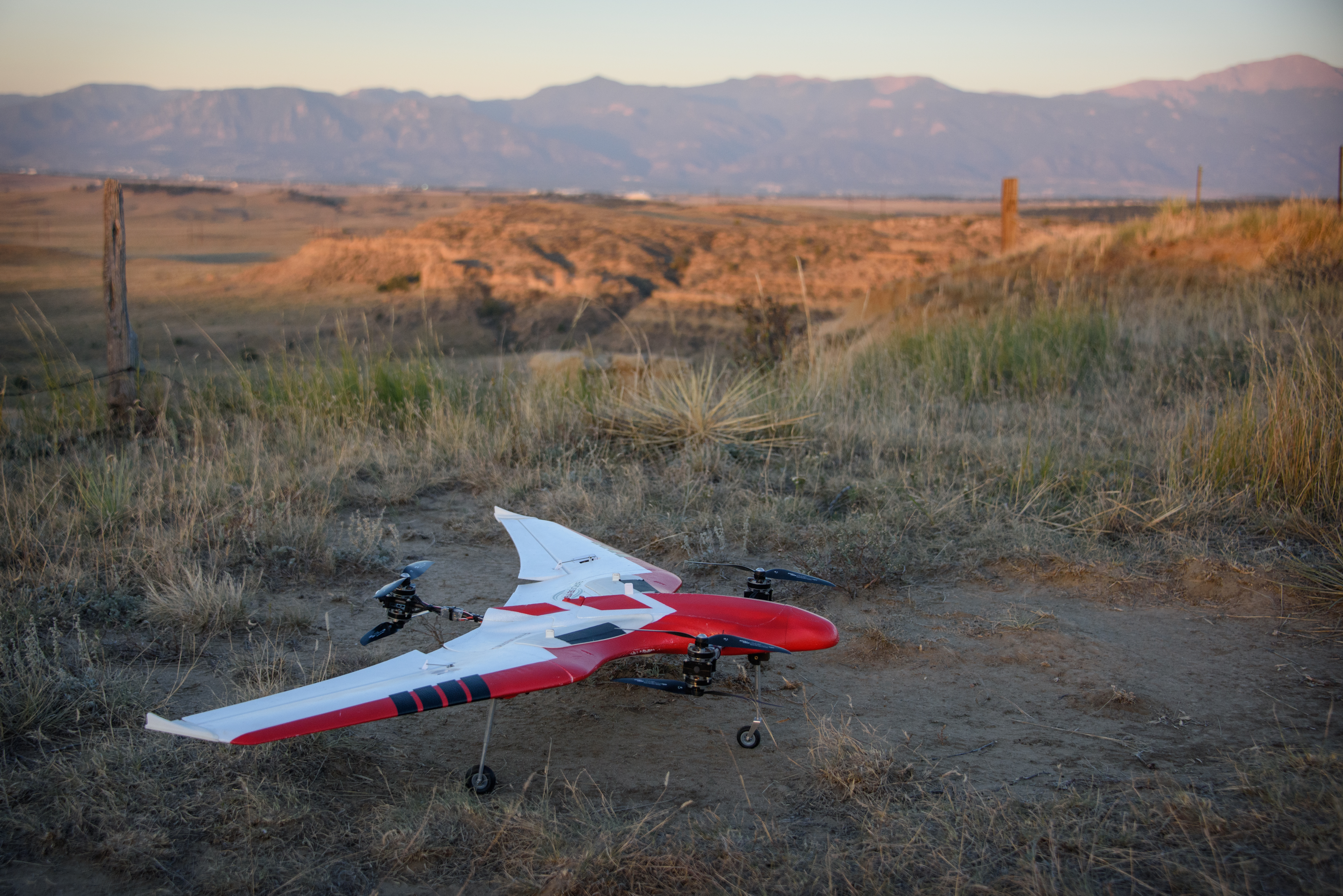 Red and white winged drone with multiple rotors rests on dry ground beside grass at Corral Bluffs, Colorado.