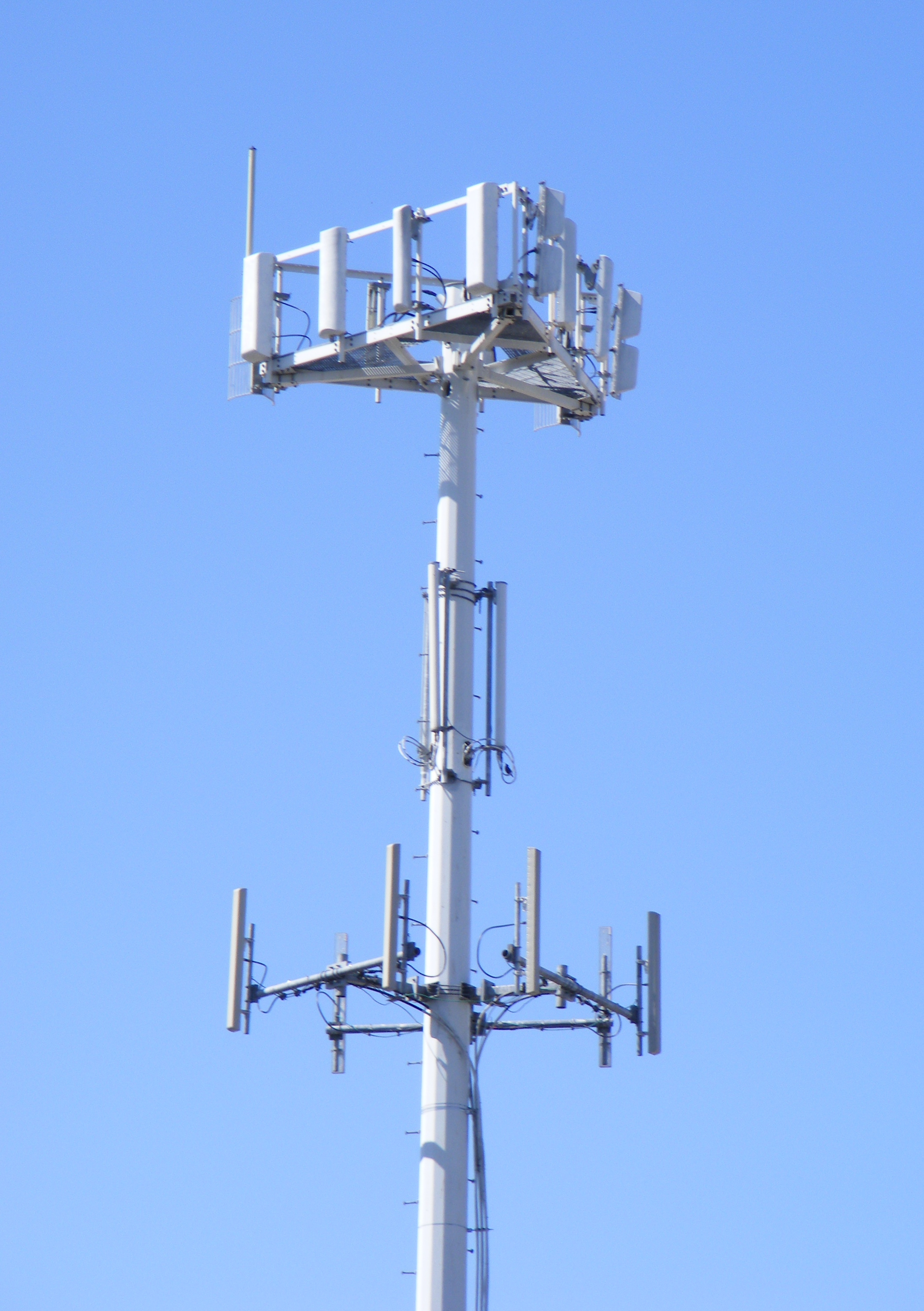 Panel antennas, supporting frames, and cables at several levels of a monopole cell tower against a blue sky.
