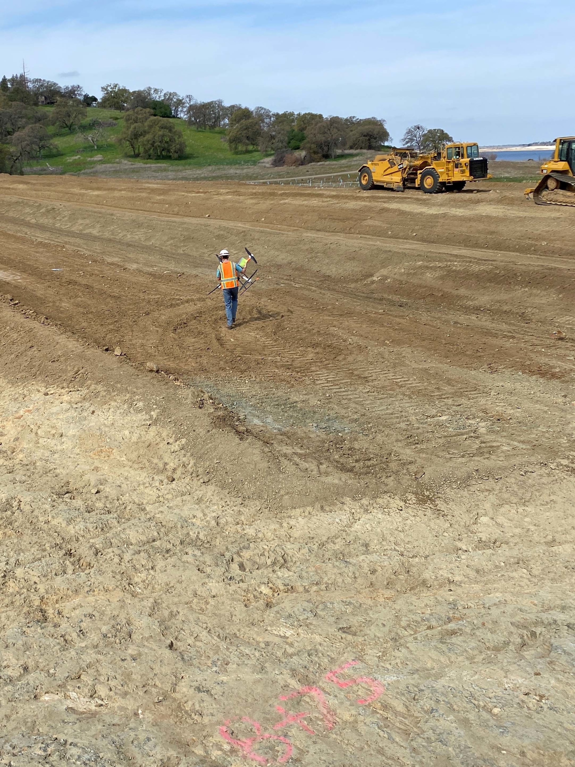 A worker carries a multirotor drone across an earthworks site, with construction machinery and Folsom Lake in the background.