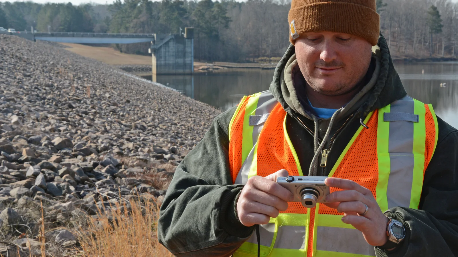 An engineer in a high-visibility vest reviews a compact camera beside the riprap slope and water at a dam.