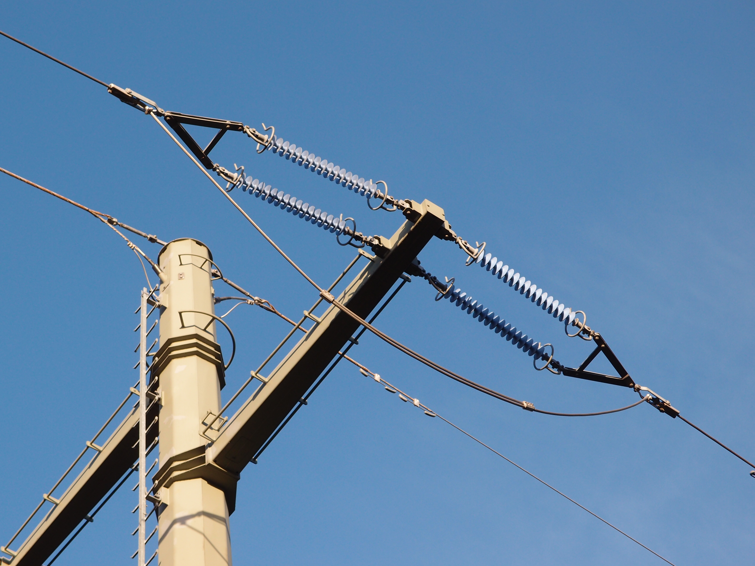 Paired ribbed insulators, metal fittings, and overhead conductors attached to a steel power-line crossarm against blue sky.