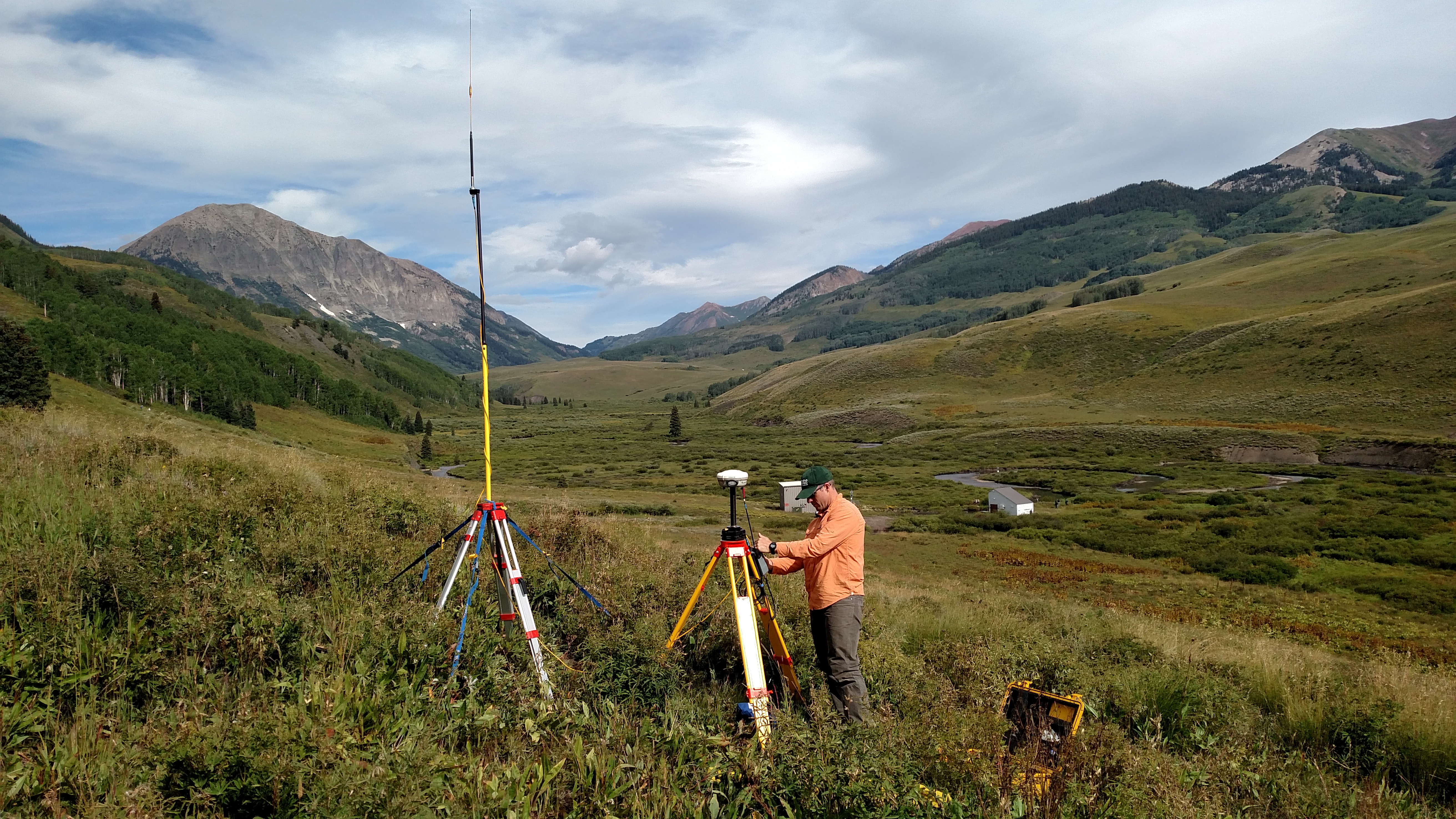 USGS scientist adjusting tripod-mounted GNSS equipment beside a radio antenna in a mountain valley