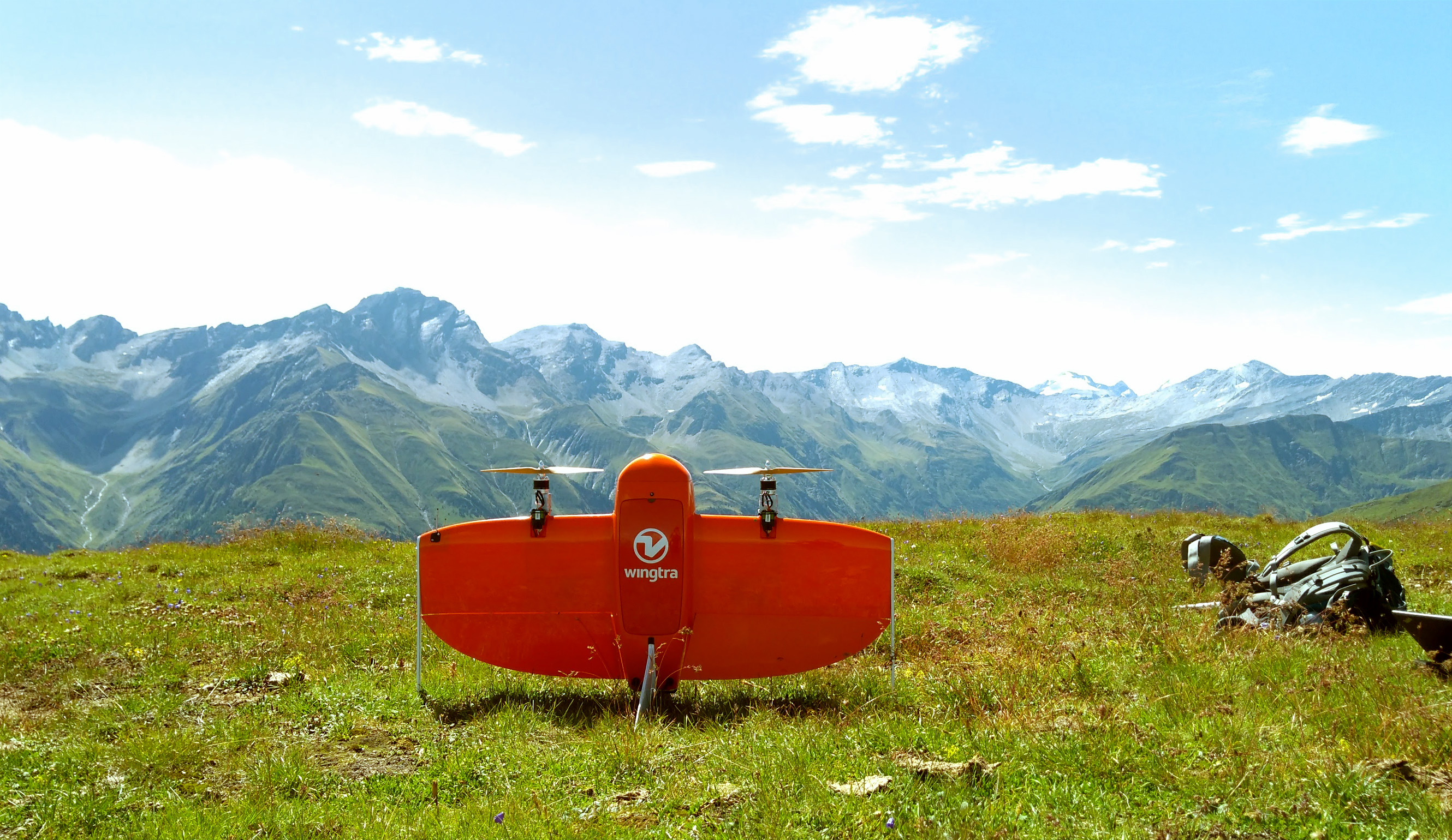 An orange WingtraOne stands upright on grass with mountains behind it.