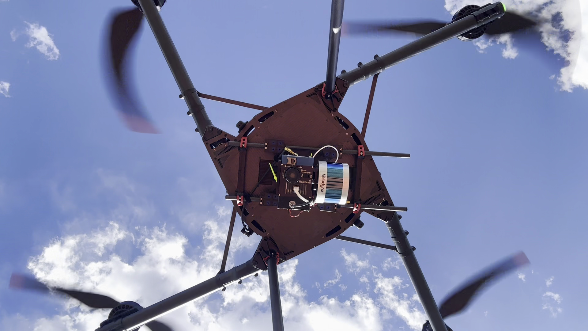 Underside of a flying multirotor showing a mounted hyperspectral camera, adjacent lidar sensor, and connecting cables.