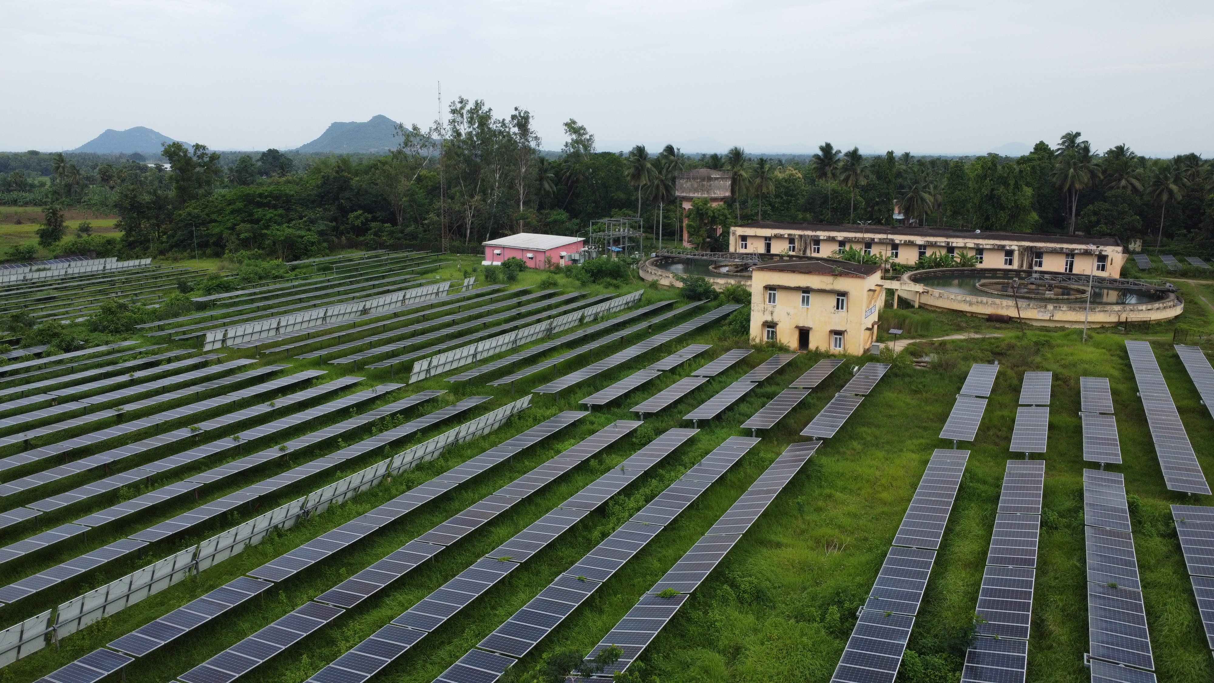 Oblique aerial view of parallel solar-panel rows beside water-treatment tanks and buildings