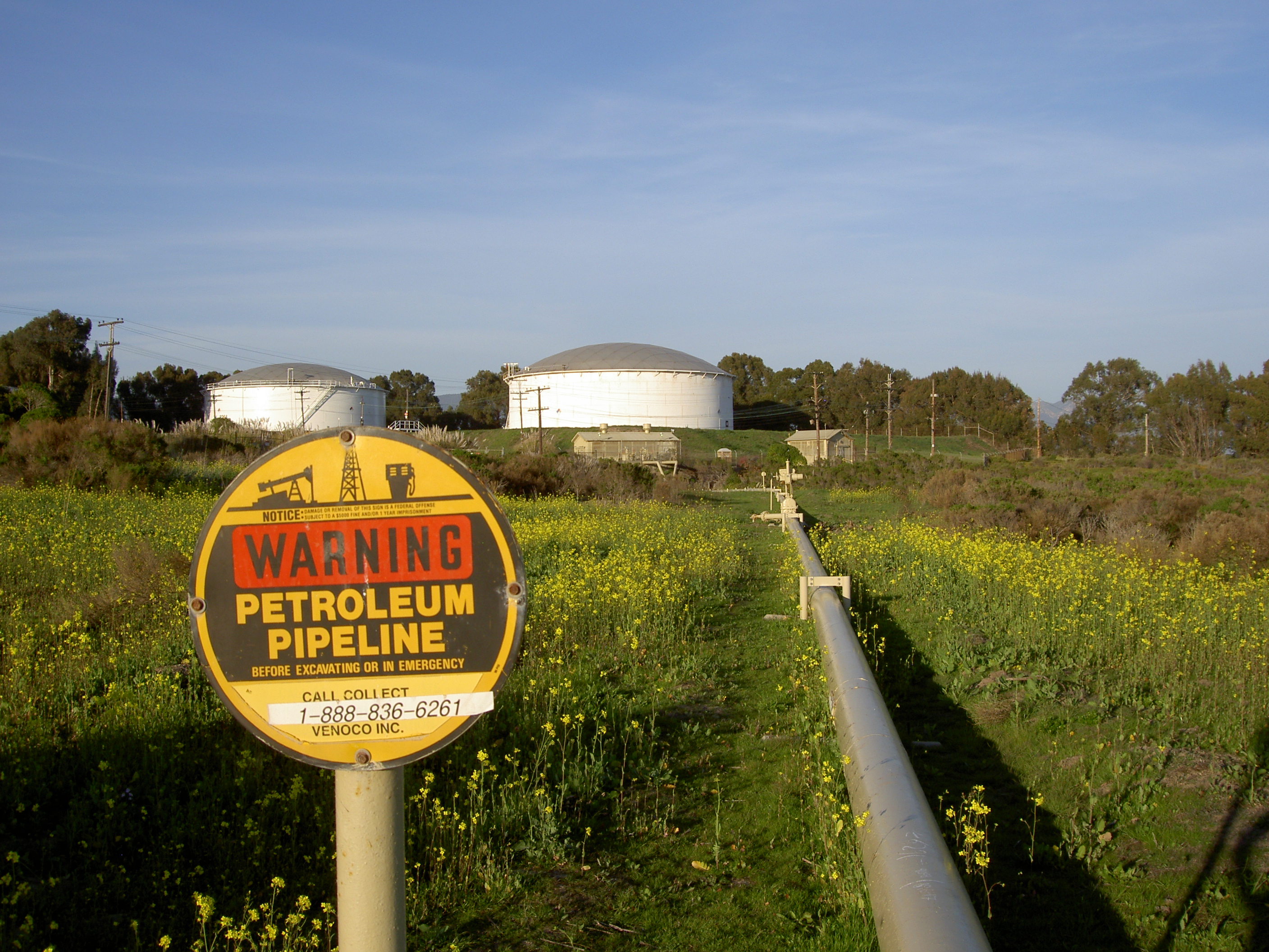 Two cylindrical storage tanks with domed roofs behind a petroleum pipeline and warning sign.