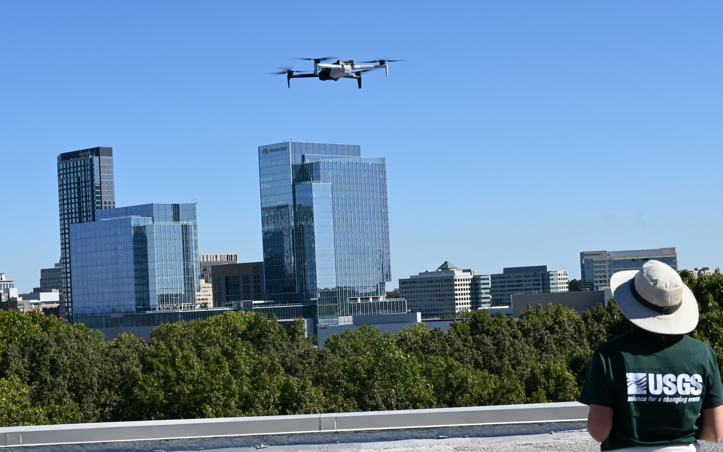 A USGS operator watches a quadcopter above a rooftop, with office buildings beyond.