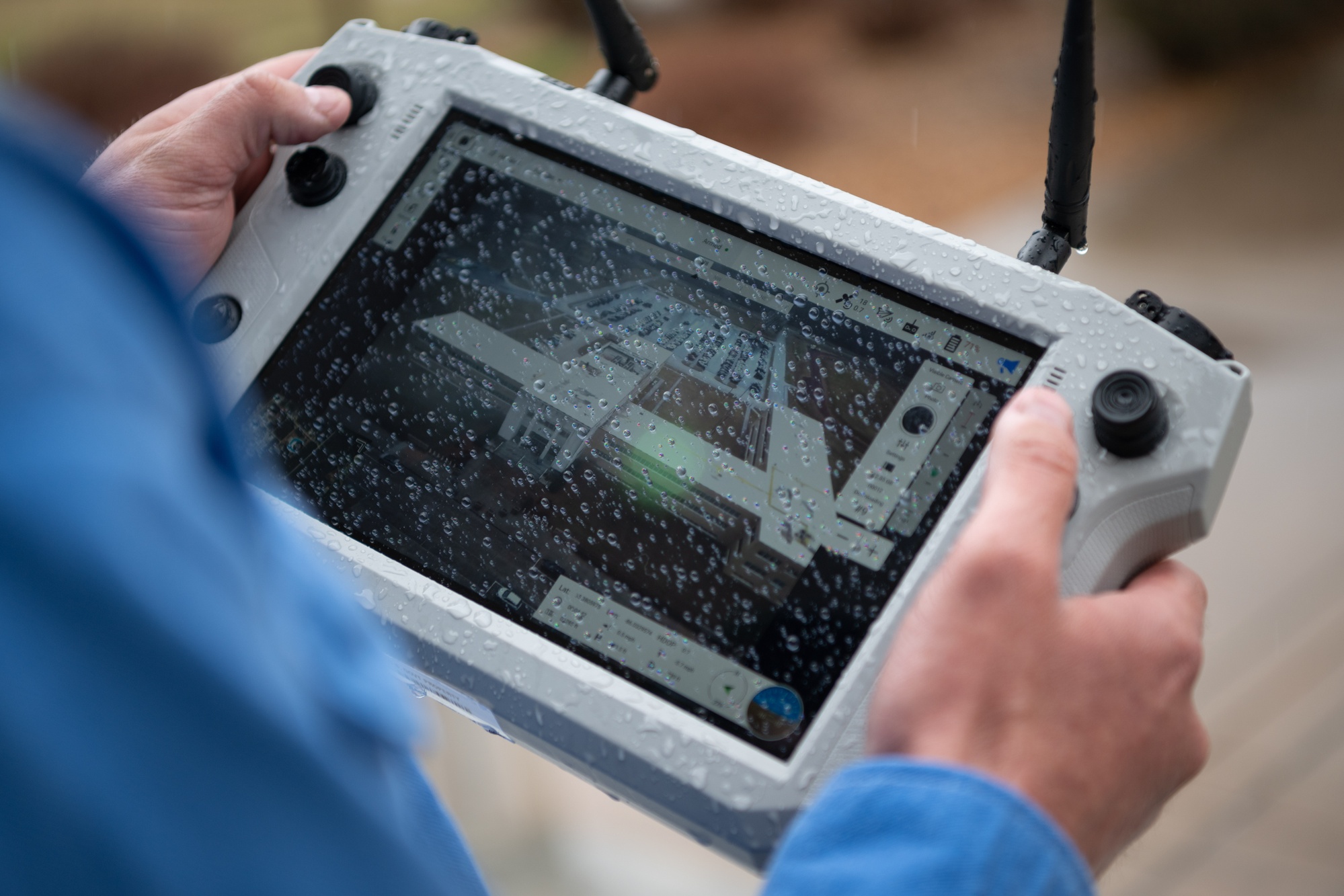 Hands holding a rain-speckled drone controller displaying an aerial view of a building and parking area