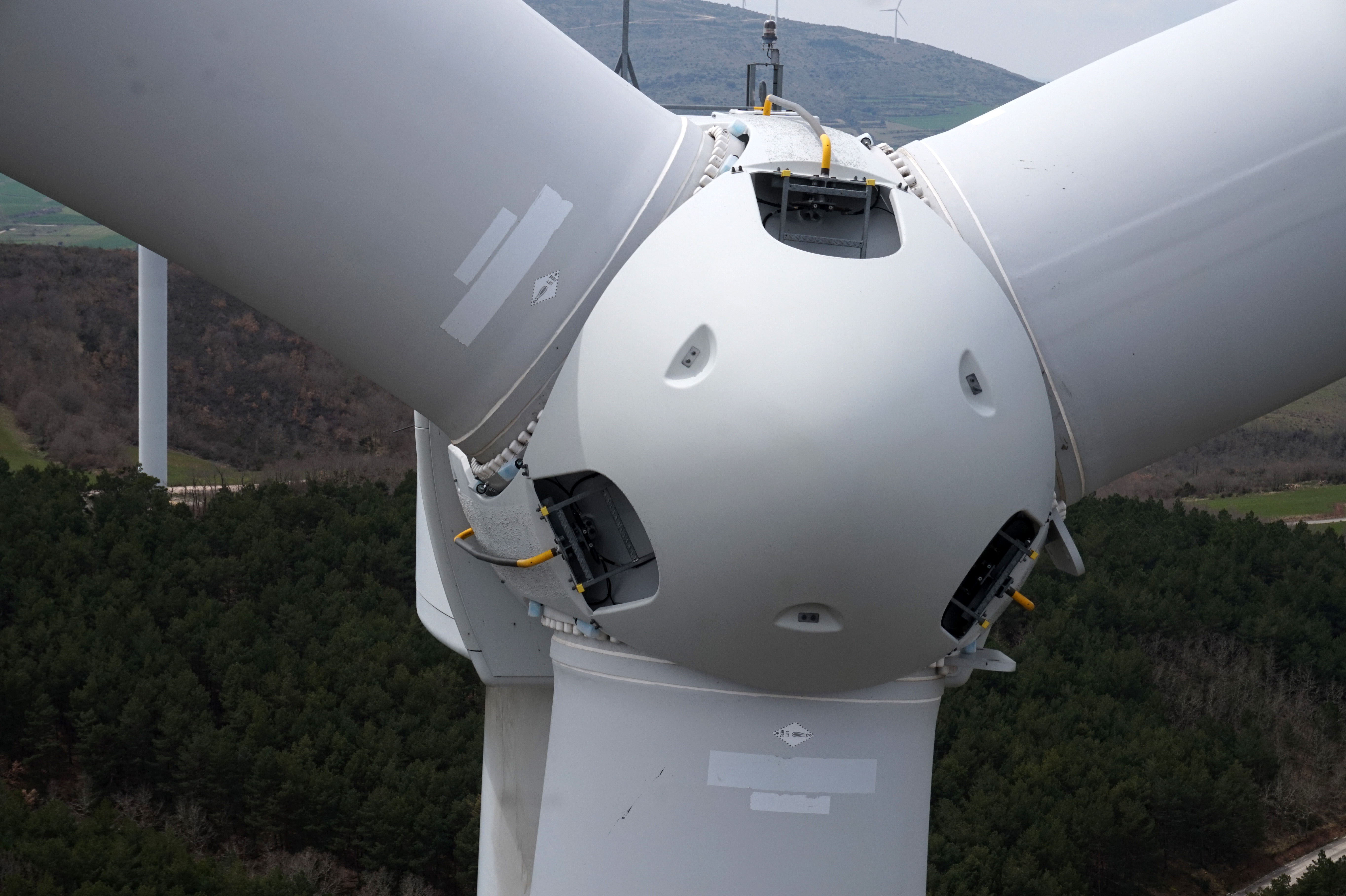 Close aerial view of a wind turbine hub, three blade roots, and open access hatches above wooded terrain.